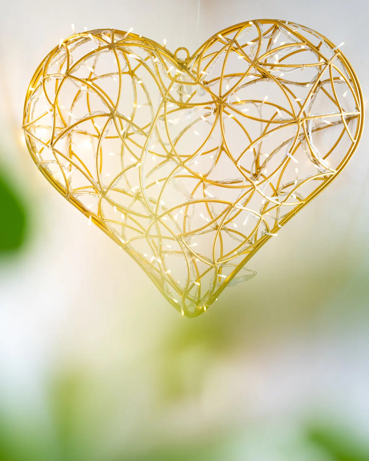 Hängendes Lichtobjekt in Herzform aus goldener Drahtstruktur – schwebend inszeniert vor weichem Hintergrund.