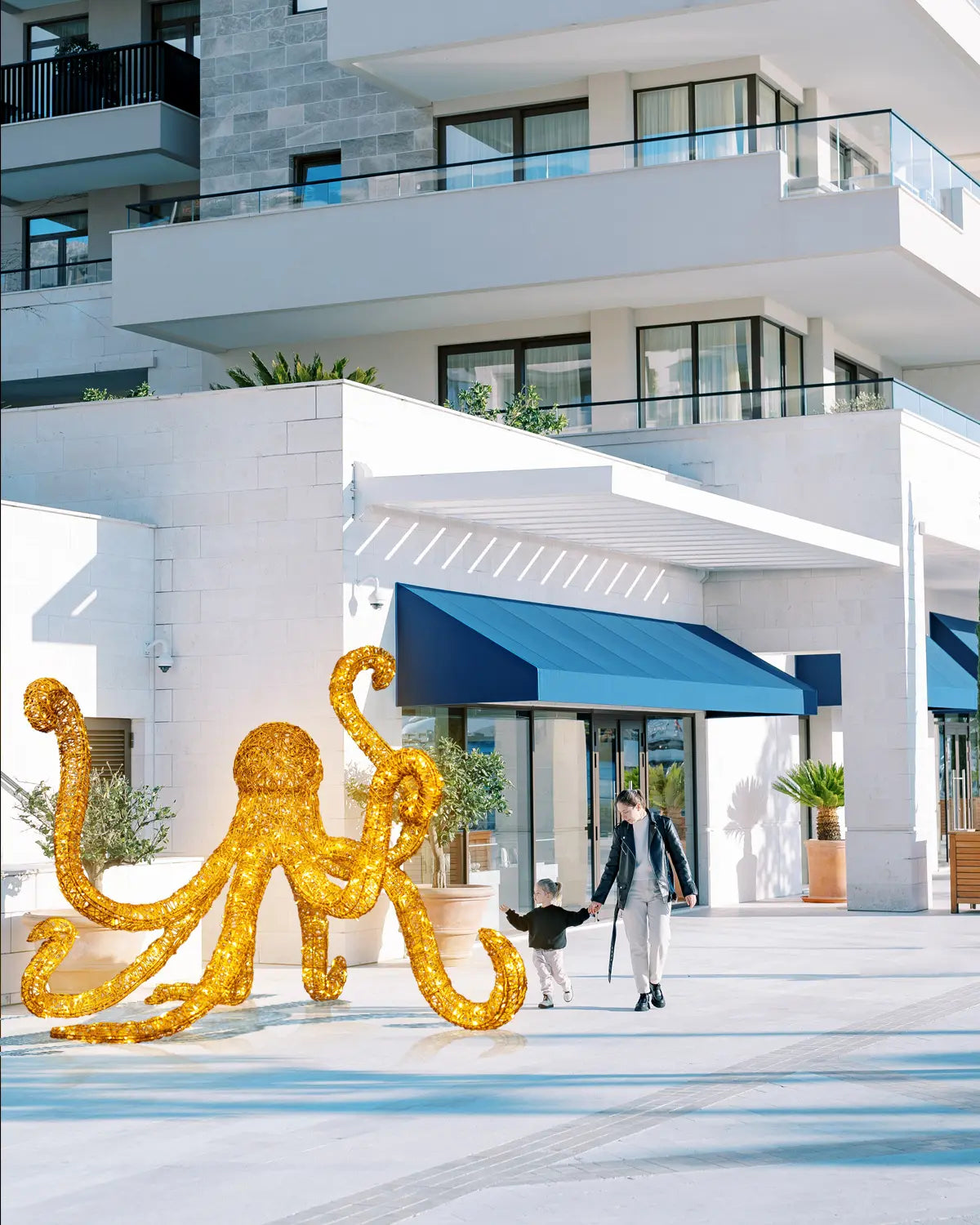 Goldene Oktopus-Lichtskulptur im Eingangsbereich eines modernen Gebäudes – Tagesszene mit Kind und Mutter beim Vorübergehen.