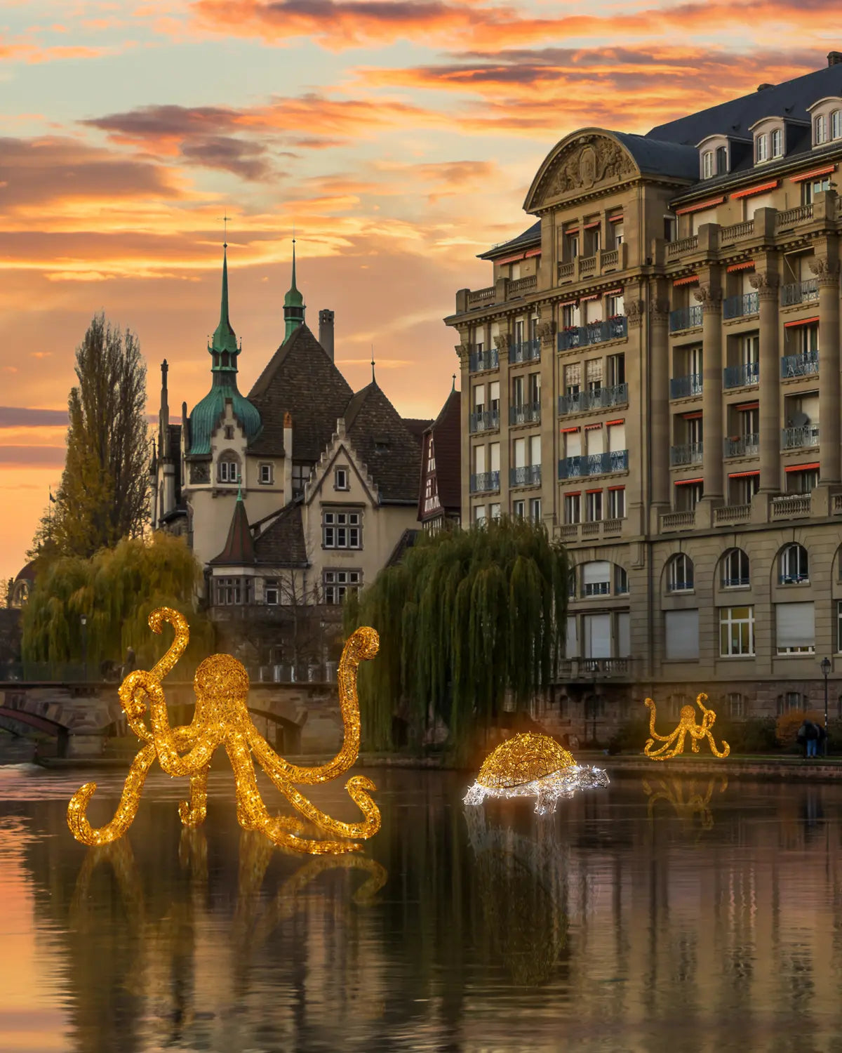 Leuchtende Oktopus- und Schildkrötenfiguren aus goldenen Lichtskulpturen schwimmen bei Sonnenuntergang auf einem ruhigen Stadtfluss, im Hintergrund historische Gebäude mit Türmen und Fassaden.