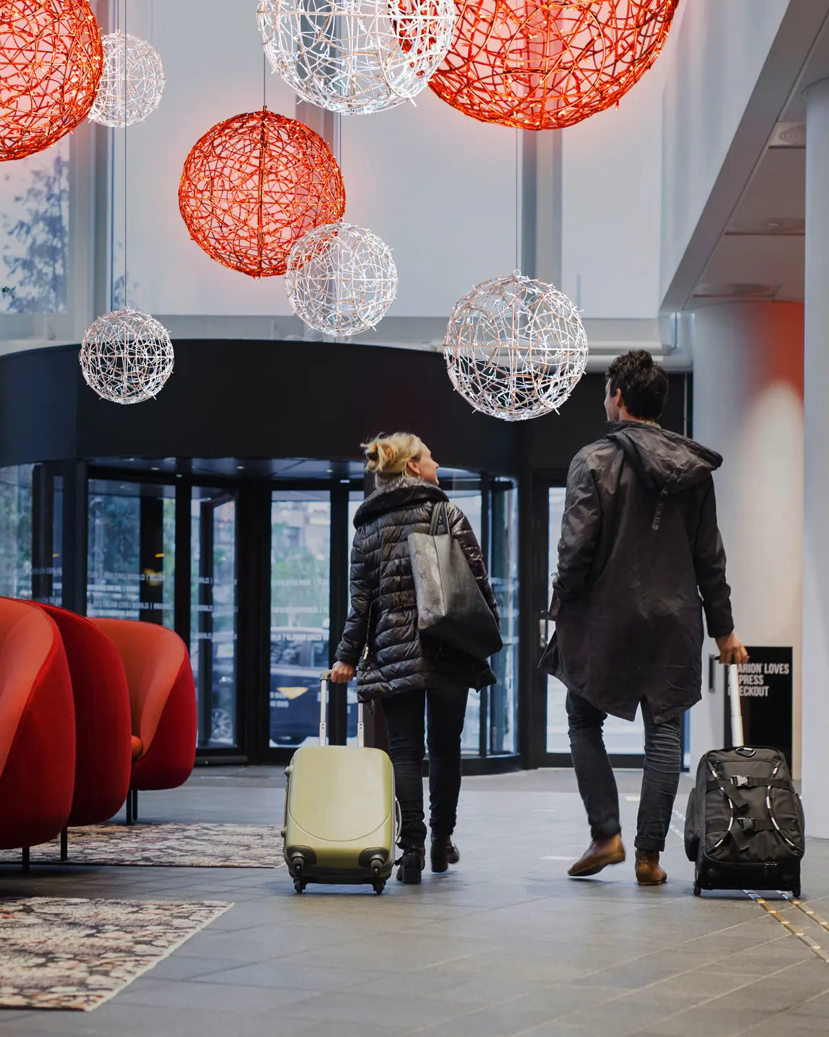Weiße und rote Lichtkugeln aus Draht als Deckeninstallation in einem modernen Hoteleingang – Gäste mit Gepäck beim Betreten der Lobby.