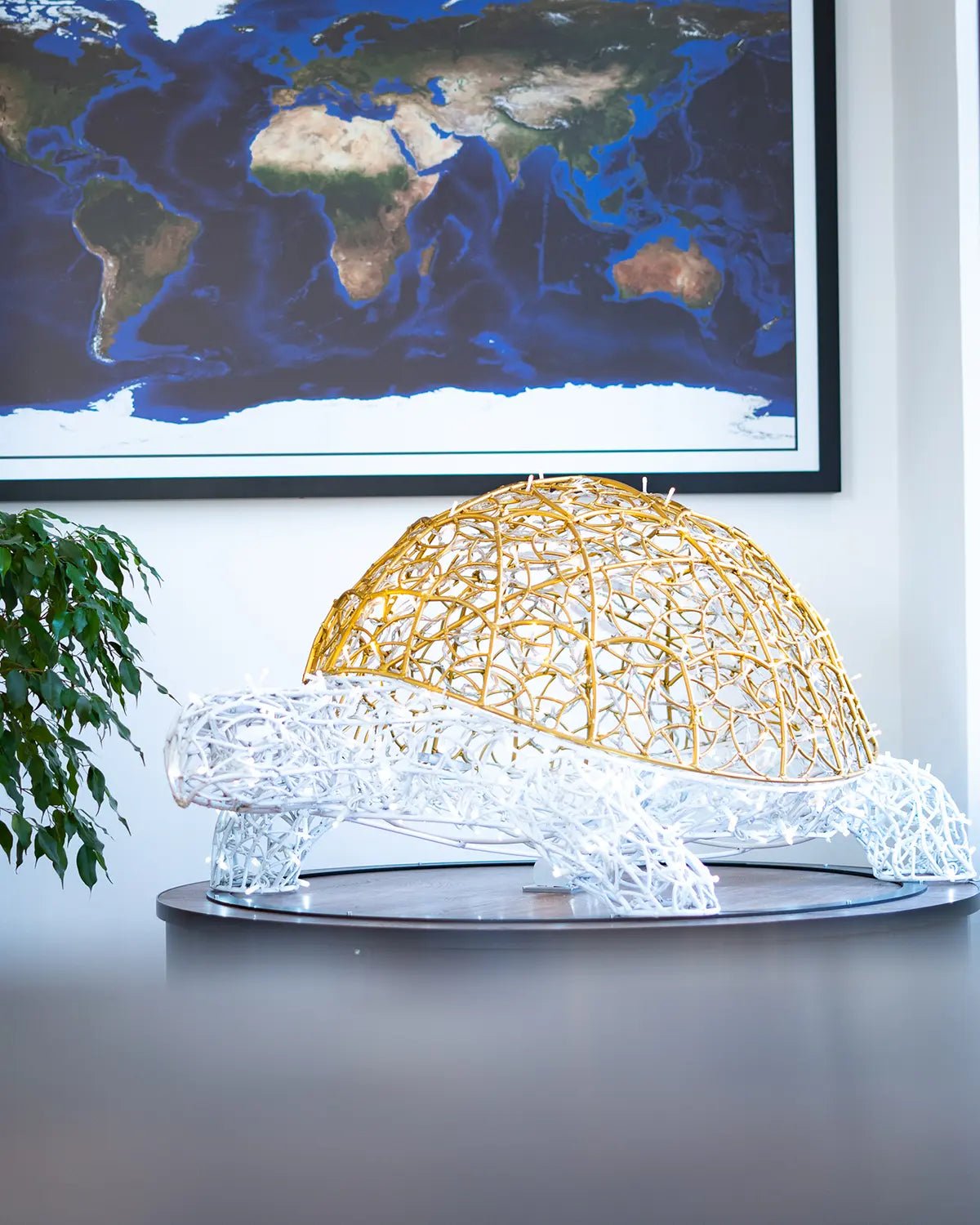 Große Lichtskulptur in Schildkrötenform aus gold-weißer Aluminiumstruktur – inszeniert im Innenraum vor Weltkarte.