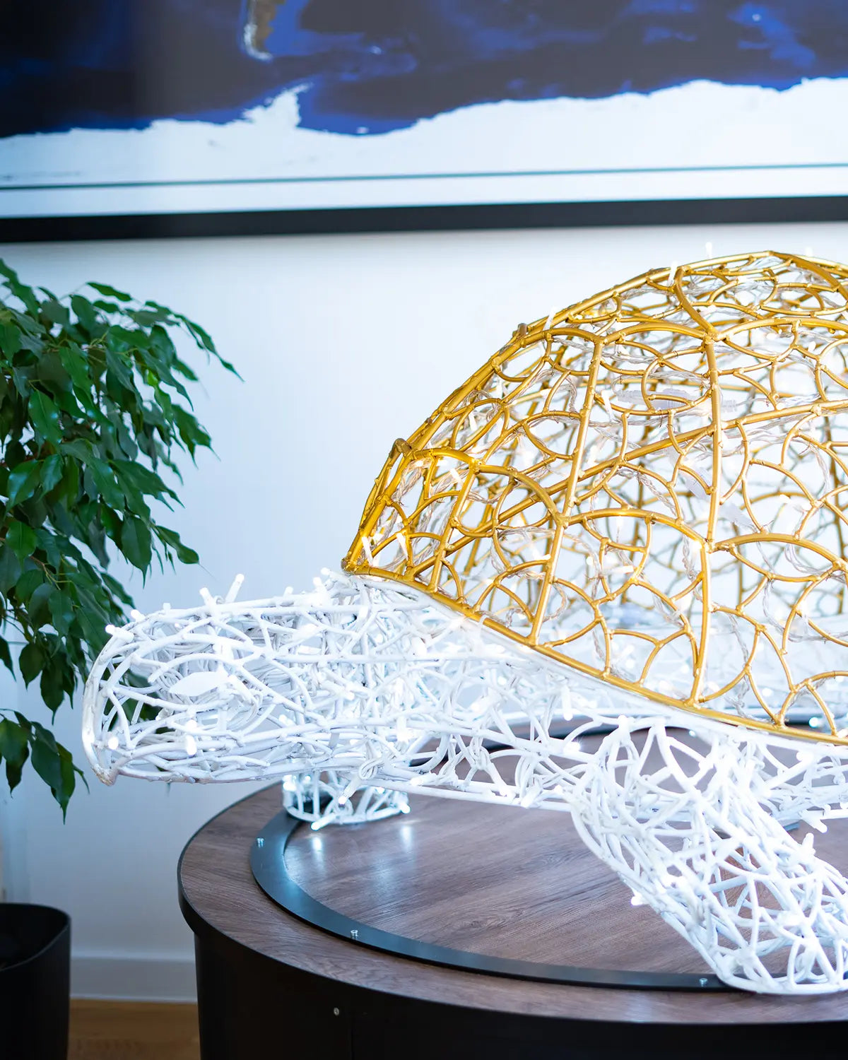 Lichtskulptur einer Schildkröte mit goldfarbenem Panzer und weißem Körper aus Draht – ausdrucksstark platziert auf rundem Holztisch im Innenraum.
