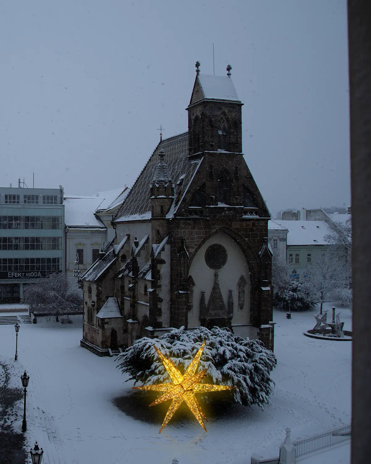 Leuchtender, goldener Achtzack-Stern aus Lichterdraht steht vor einer verschneiten historischen Kirche auf einem Platz. Die Szene spielt sich bei dichtem Schneefall und gedämpftem Tageslicht ab, wodurch die Lichter des Sterns besonders hervorstechen.