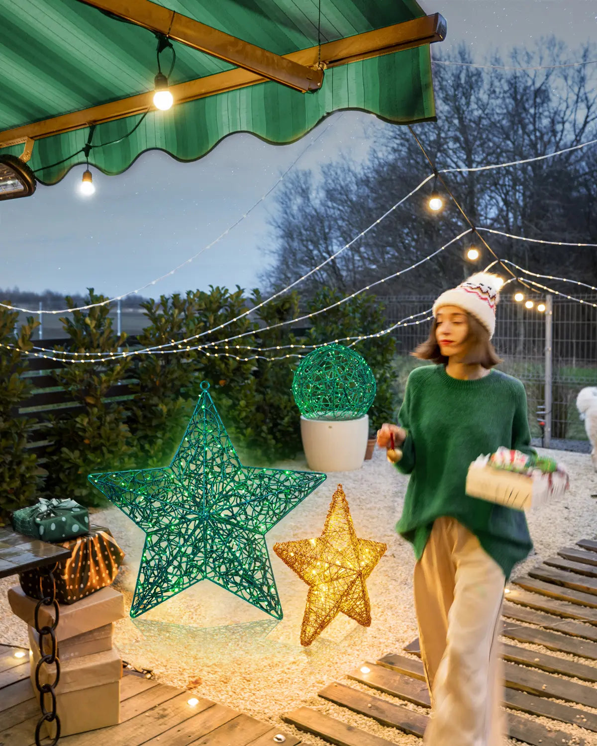 Frau mit Geschenken auf winterlicher Terrasse neben grüner und goldener Alganic-Leuchtstern-Skulptur – Outdoor-Weihnachtsdeko im Gartenambiente mit Lichterketten.