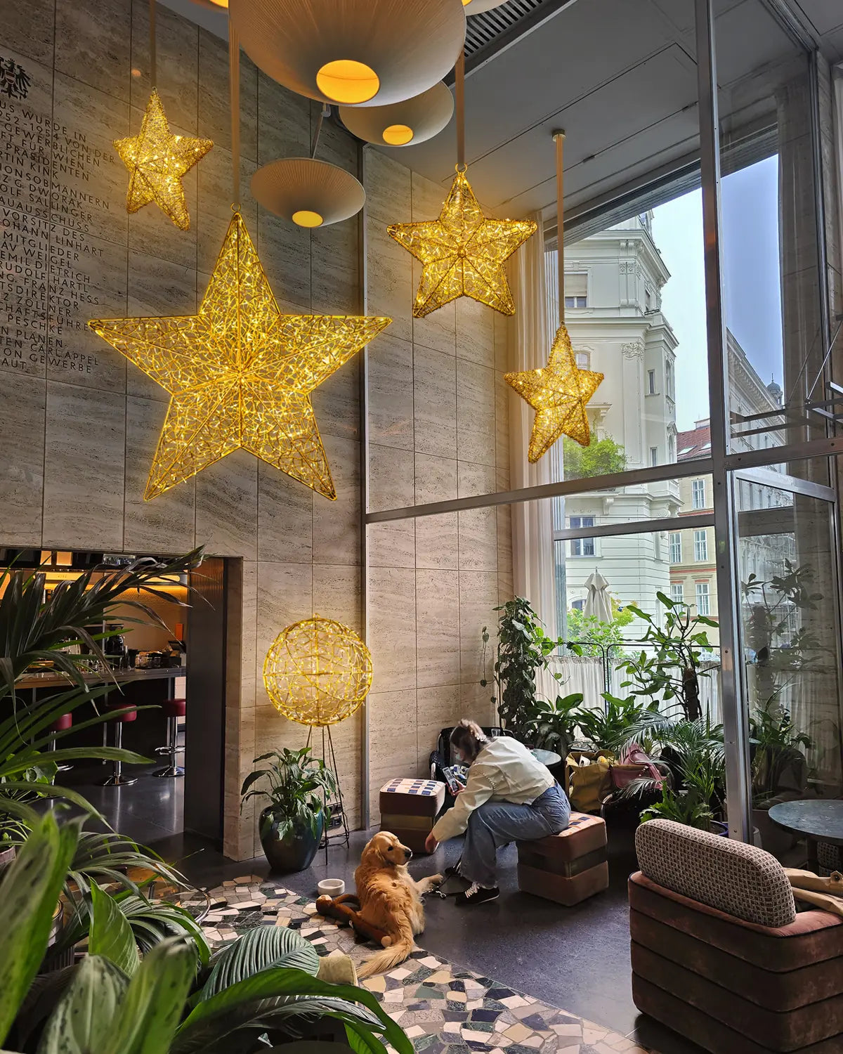 Mehrere goldene Sterne hängen als Deckenbeleuchtung in einer gemütlichen Lounge mit Pflanzen, einem Hund und einer jungen Frau.