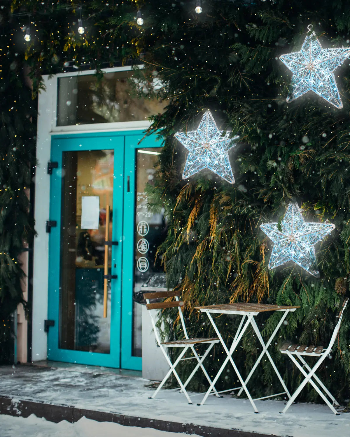Leuchtende weiße Sterne an winterlich geschmückter Fassade – Alganic-Lichtobjekte über Sitzplatz vor einem Eingangscafé.